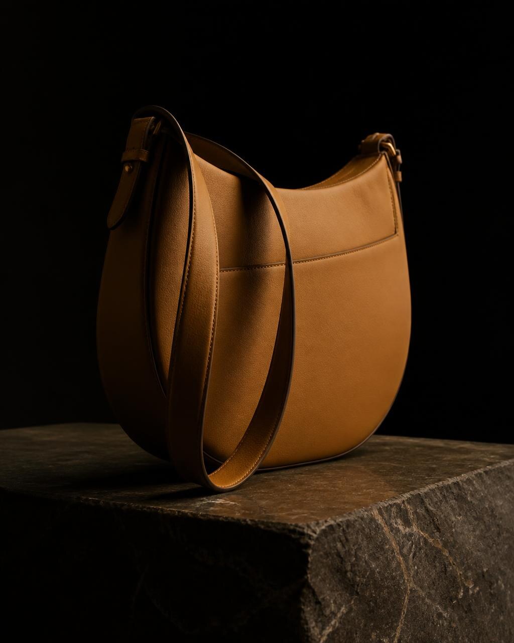 The Lindiwe half-moon shoulder bag in warm caramel nyala leather on a dark stone plinth